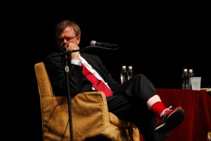 Photo of Garrison Keillor on stage sitting in an armchair in front of a microphone