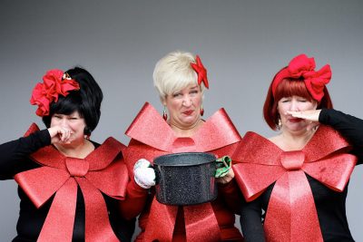 Photo by Tammy Brice, courtesy of Plymouth Playhouse. Three of the Looney performers holding a stinking pot of lutefisk and holding their noses. (They're also adorned in huge holiday bows.)