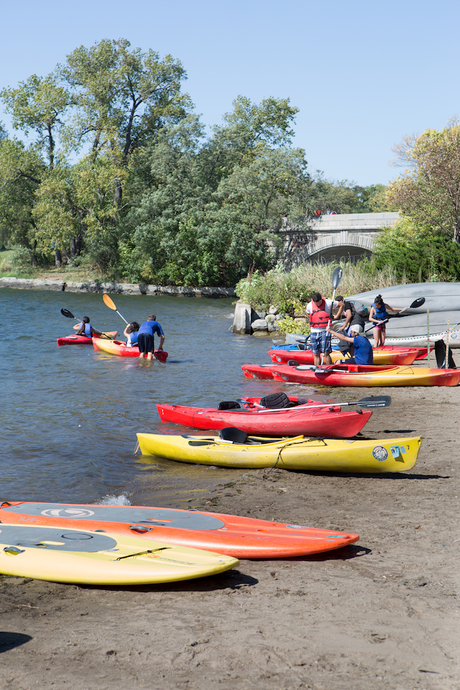 Orange and red kayaks on the shore of a lake with people in life jackets.