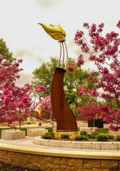 A tall, gently curving public art statue has a golden flower blossom at the end of it.