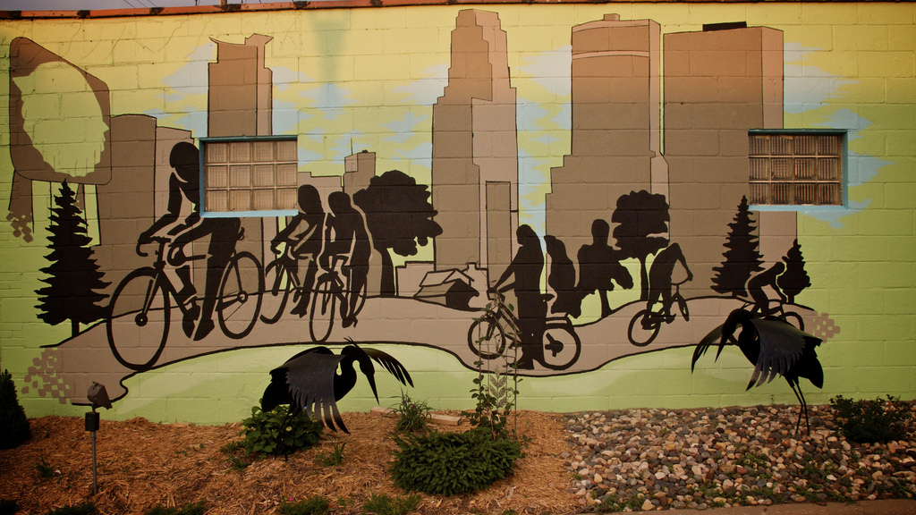 Photo by Bjorn/flickr. Bicyclists on a city path with the Minneapolis skyline in the background. A nice 3D touch is added with the metal birds in front of it.