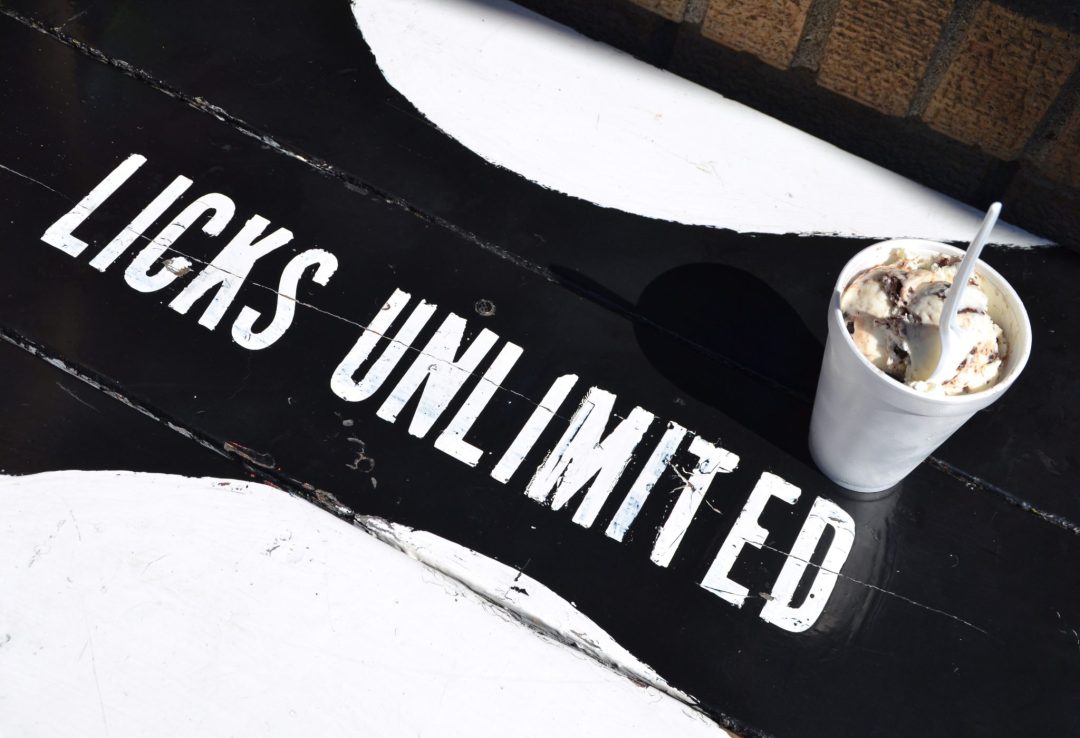 A styrofoam cup filled with ice cream sitting on a table with a black and white Licks Unlimited logo painted on it.