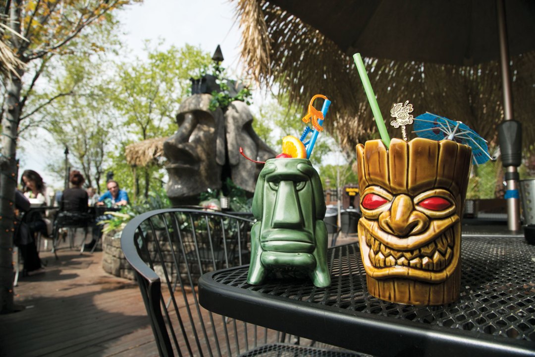 Two tiki-inspired mugs are sitting on a table on Psycho Suzi's patio. Easter Island-inspired heads are sitting in the background.